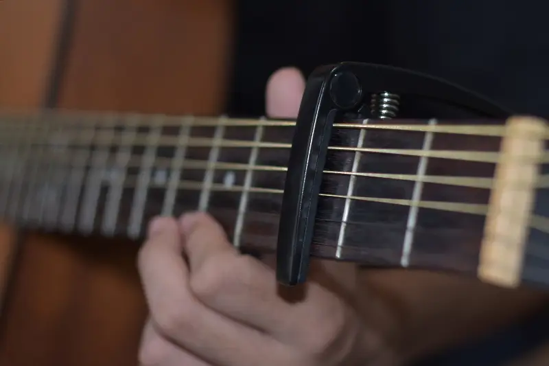 Capodastre à pince noir serré sur le manche d'une guitare folk, devant la main gauche du guitariste