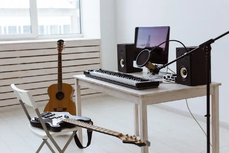 Petit bureau blanc avec clavier maître, micro à condensateur sur pied, deux enceintes et un écran, une guitare folk et une guitare électrique à côté