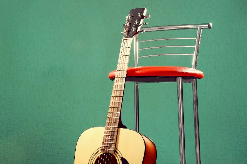 Guitare acoustique appuyée contre un tabouret de bar à l'assise rouge, devant un mur vert