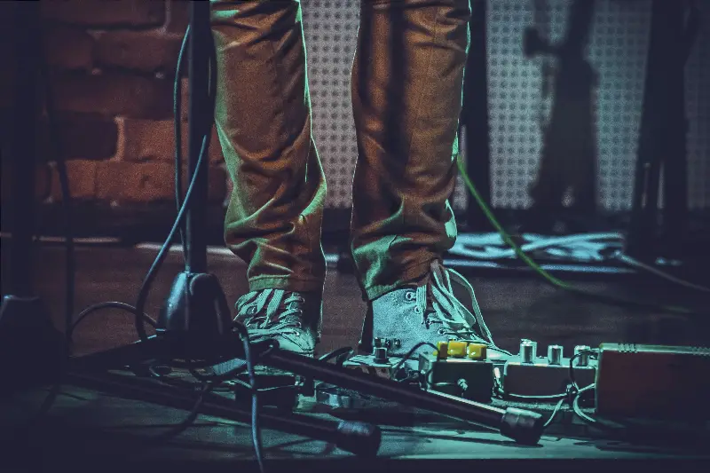Guitariste en baskets debout sur scène derrière un pédalier d'effets, pied de micro au premier plan