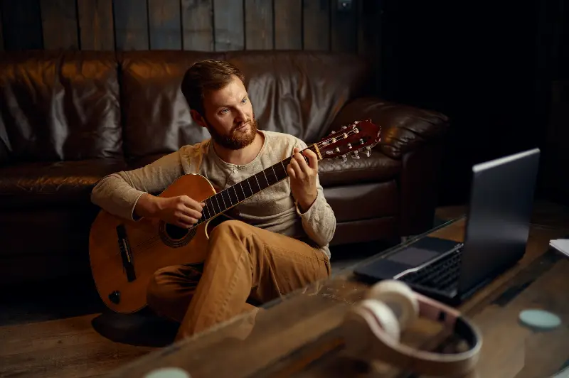 Homme assis par terre devant un canapé en cuir, qui joue de la guitare classique, un ordinateur portable et un casque sur la table basse