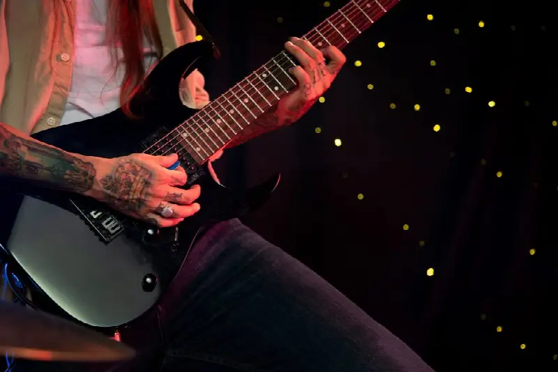 Guitariste aux bras tatoués jouant d'une guitare électrique noire devant un fond sombre parsemé de points lumineux