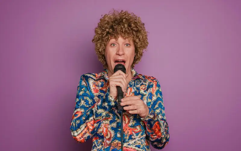Un chanteur en perruque frisée et chemise à motifs chante dans un micro devant un fond violet