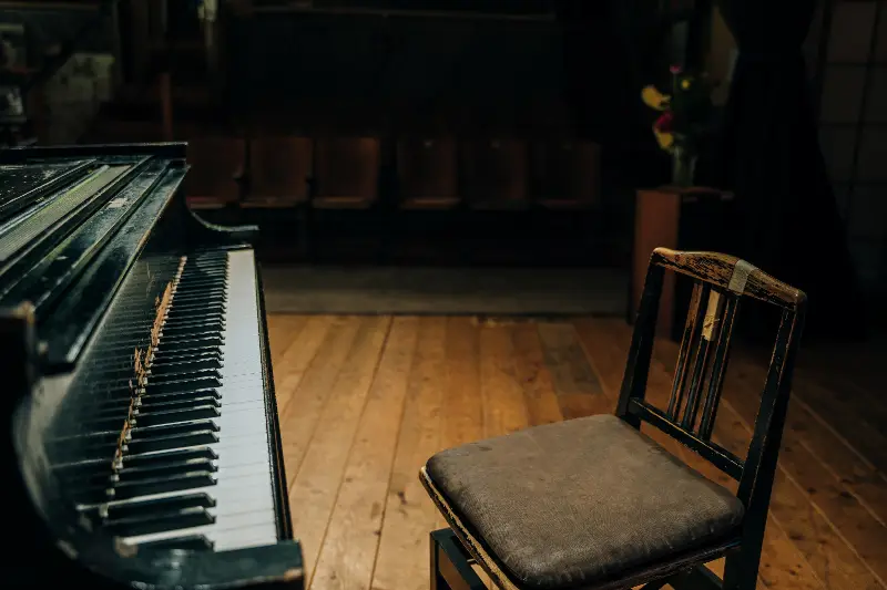 Vieux piano à queue noir aux touches usées et sa chaise de pianiste sur un plancher, devant des rangées de sièges